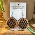 Pair of round wooden drop earrings with a fish-scale cutout pattern, hanging from silver hooks on a white display card from Willis Design Co., set against a warm wood and ceramic background.