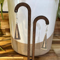 A pair of geometric drop earrings with triangular metal pendants featuring horizontal lines and a cutout, displayed on a wooden stand in front of a ceramic planter.