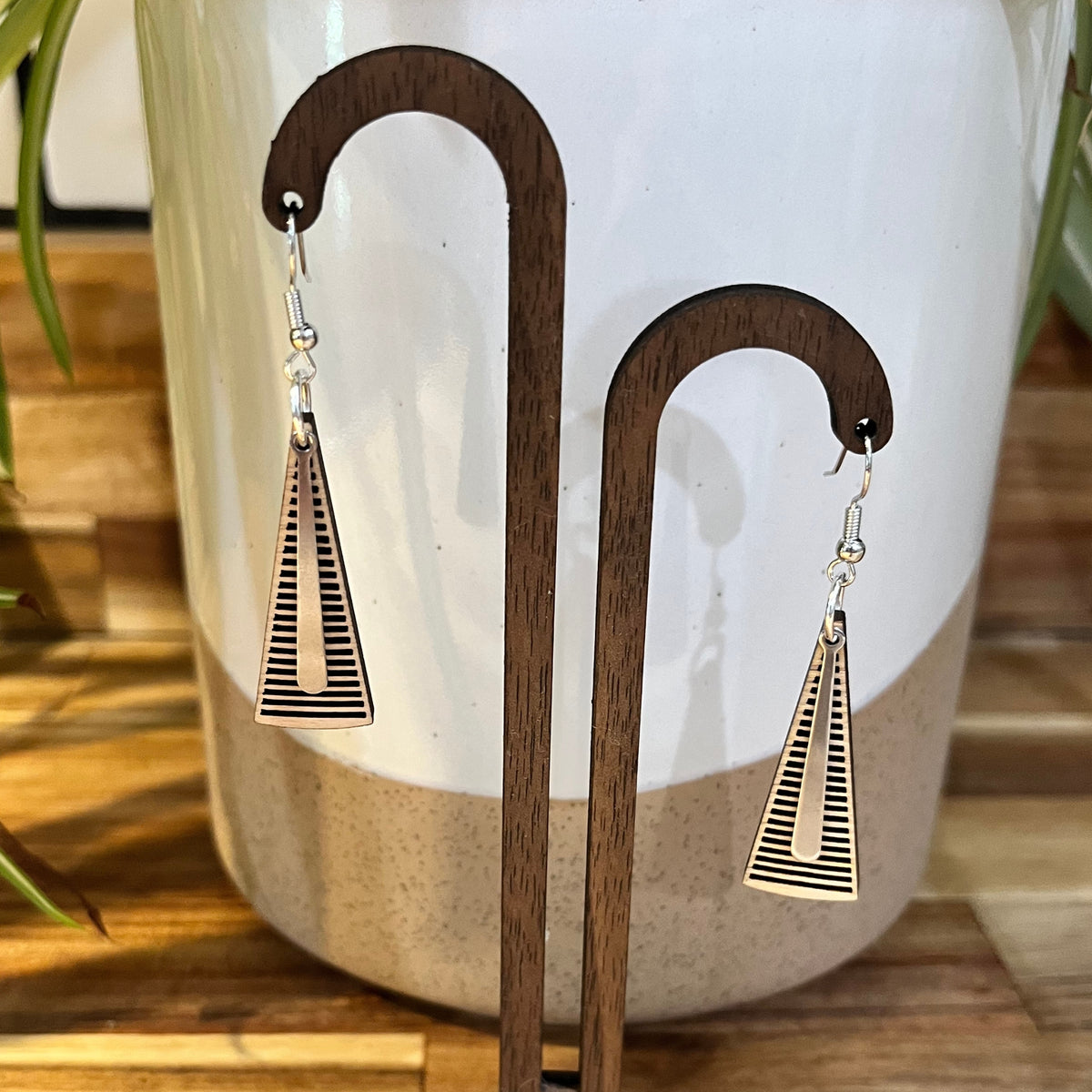 A pair of geometric drop earrings with triangular metal pendants featuring horizontal lines and a cutout, displayed on a wooden stand in front of a ceramic planter.