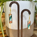 A pair of rectangular drop earrings with gold-tone hardware and colorful marbled resin inserts, displayed on a wooden stand in front of a ceramic planter with green leaves.