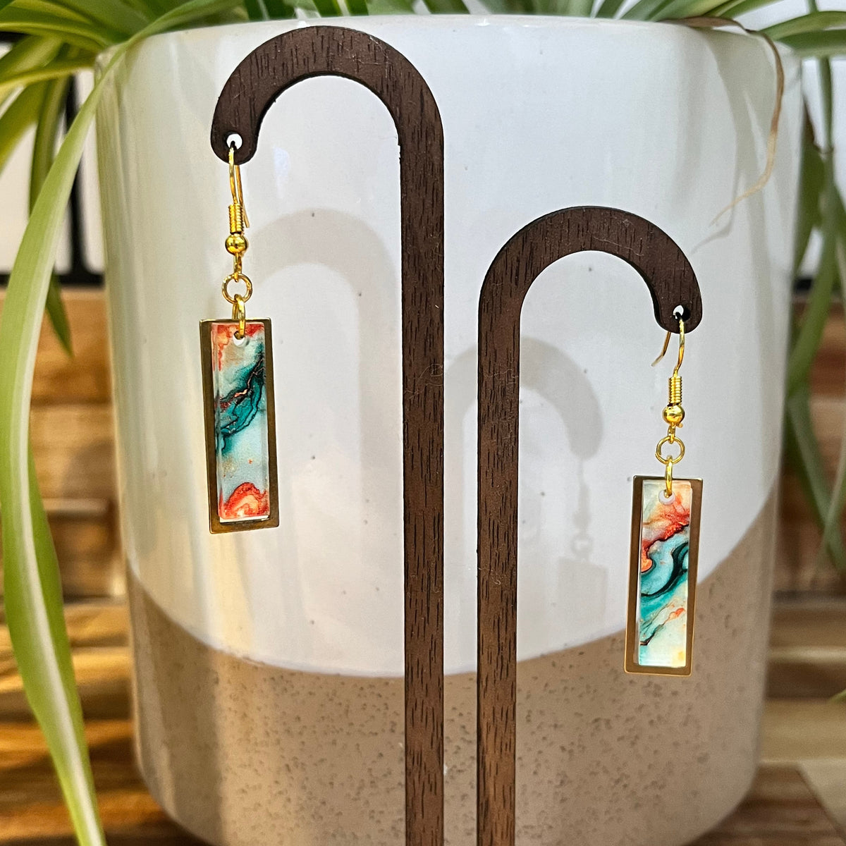 A pair of rectangular drop earrings with gold-tone hardware and colorful marbled resin inserts, displayed on a wooden stand in front of a ceramic planter with green leaves.