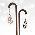 A pair of dangling earrings displayed on a wooden stand against a soft white background with blurred light bokeh. The earrings have a light pink base with black and white geometric triangle patterns and small gold dots.