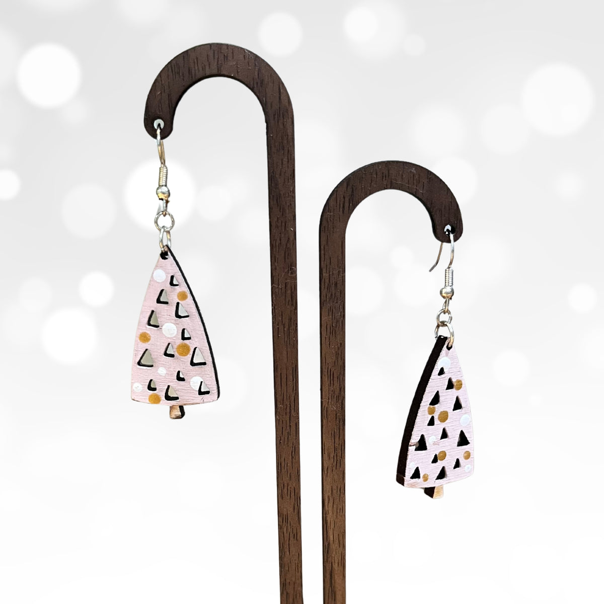 A pair of dangling earrings displayed on a wooden stand against a soft white background with blurred light bokeh. The earrings have a light pink base with black and white geometric triangle patterns and small gold dots.