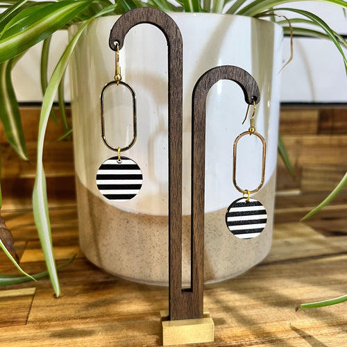 Black and white striped dangle earrings with gold-tone hardware, displayed on a wooden stand in front of a white ceramic planter with green plant leaves.