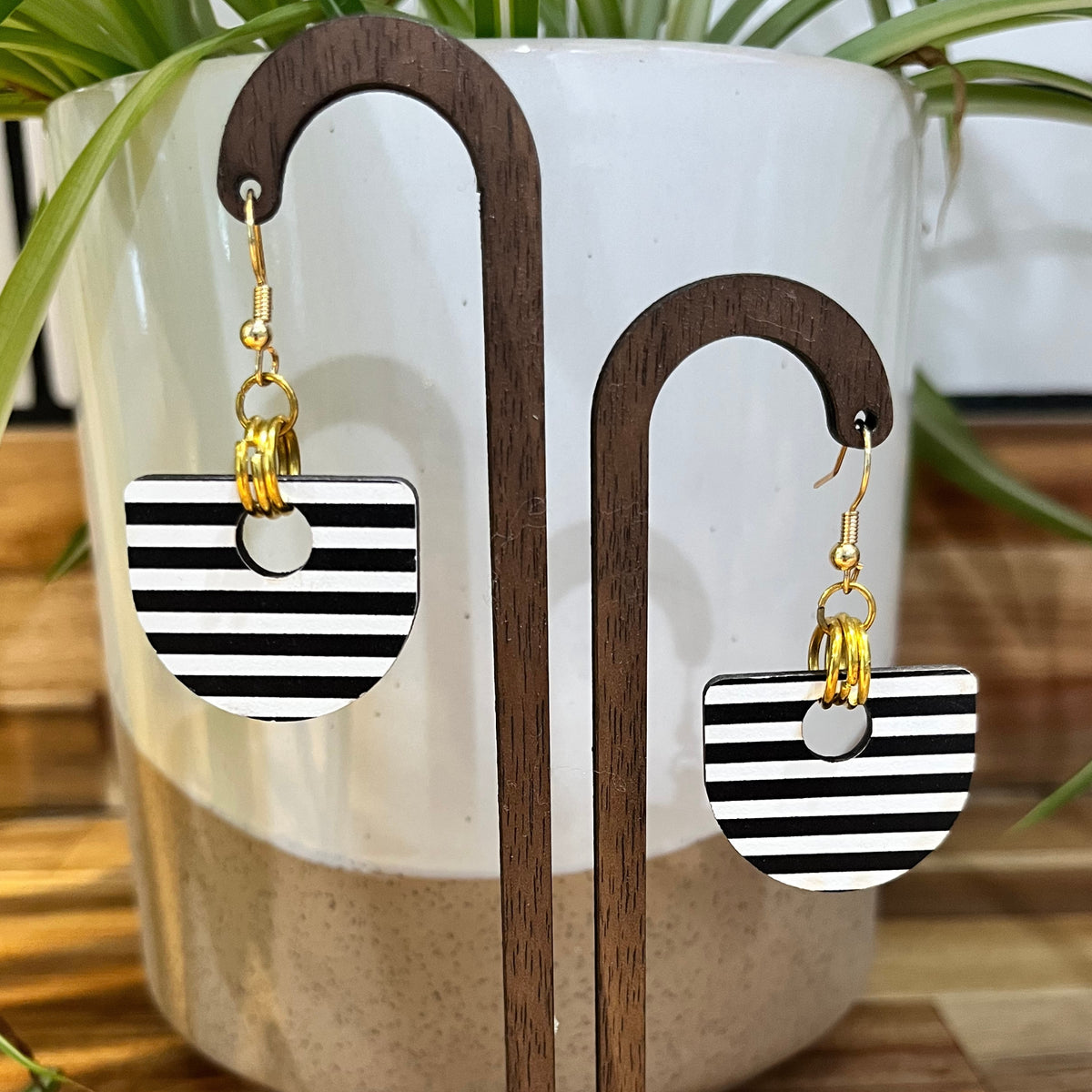 Black and white striped semi-circular drop earrings with gold hooks, displayed on a wooden stand in front of a white planter with green leaves.