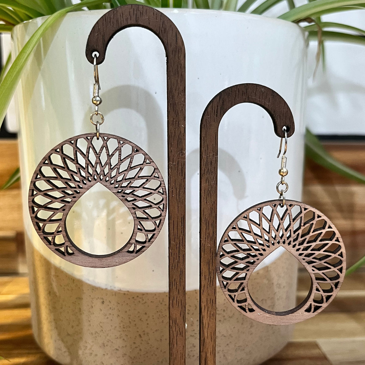 A pair of laser-cut wooden dangle earrings with a teardrop-shaped cutout and intricate geometric web patterns, displayed on a wooden stand in front of a white ceramic pot with green leaves.