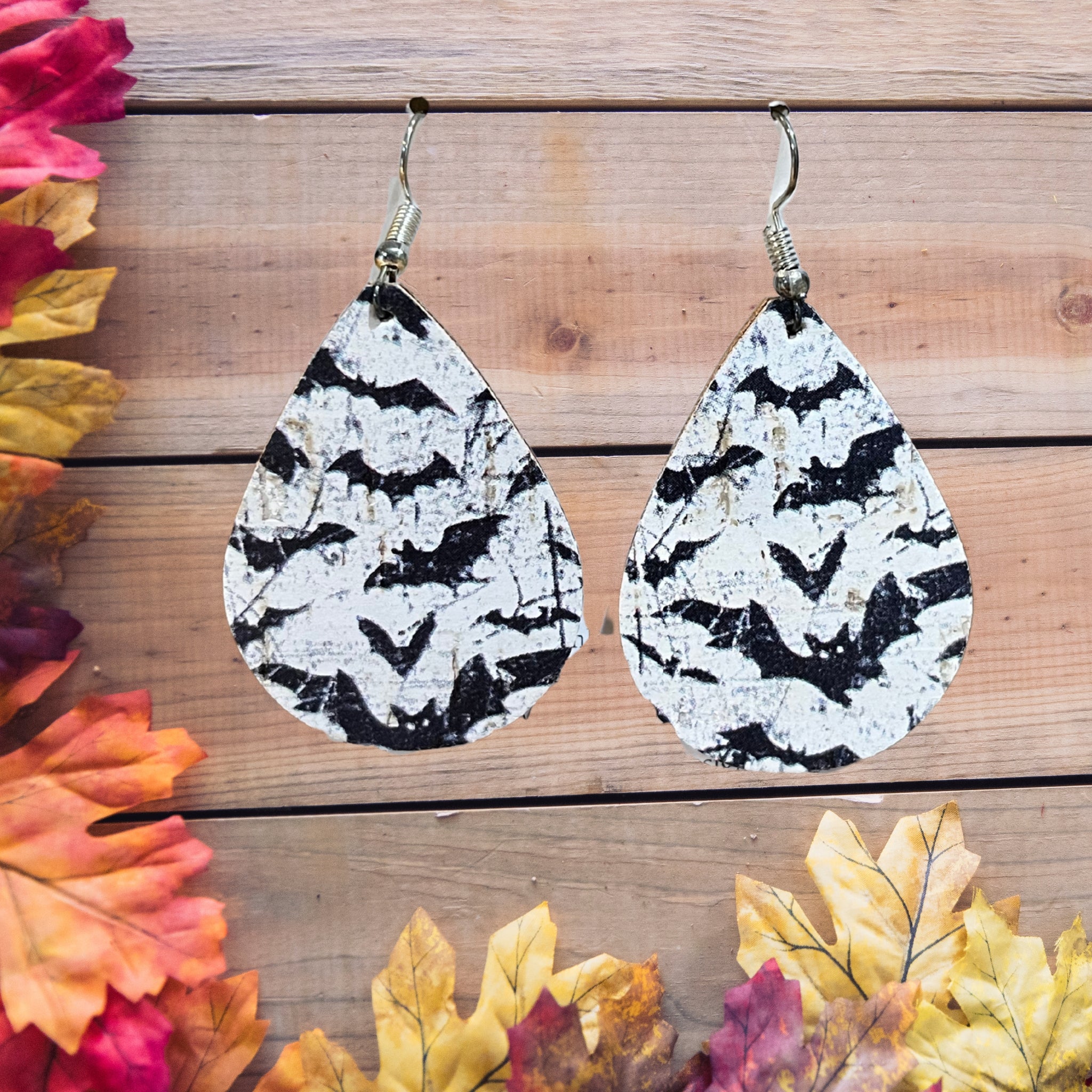 A pair of teardrop-shaped dangle earrings with a black and white bat pattern, displayed on a rustic wooden background with autumn leaves.
