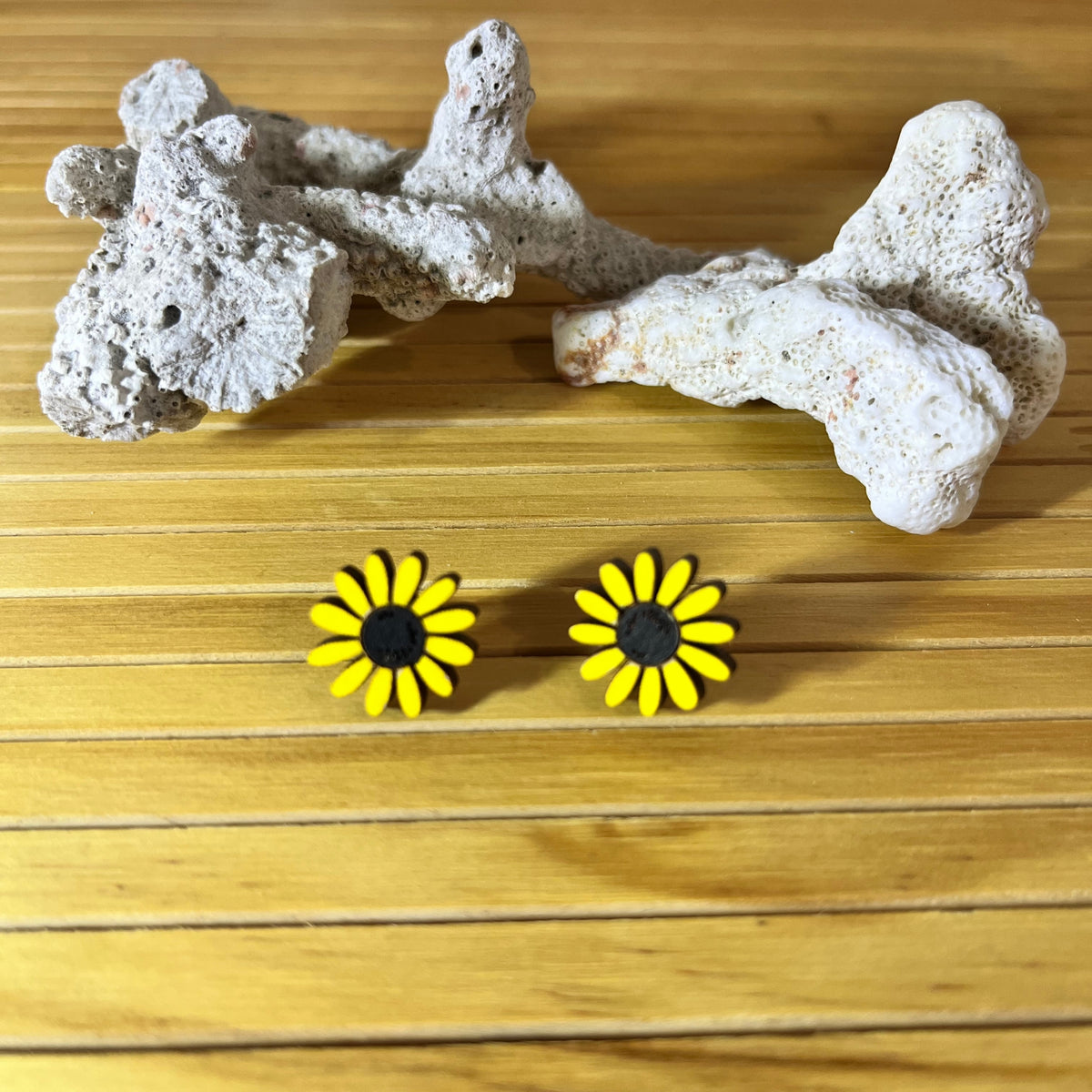 A pair of yellow daisy stud earrings with black centers, displayed on a light wood surface below a piece of white coral.