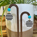 Black and white striped dangle earrings with turquoise blue round studs, displayed on a wooden stand in front of a white ceramic planter with green leaves.