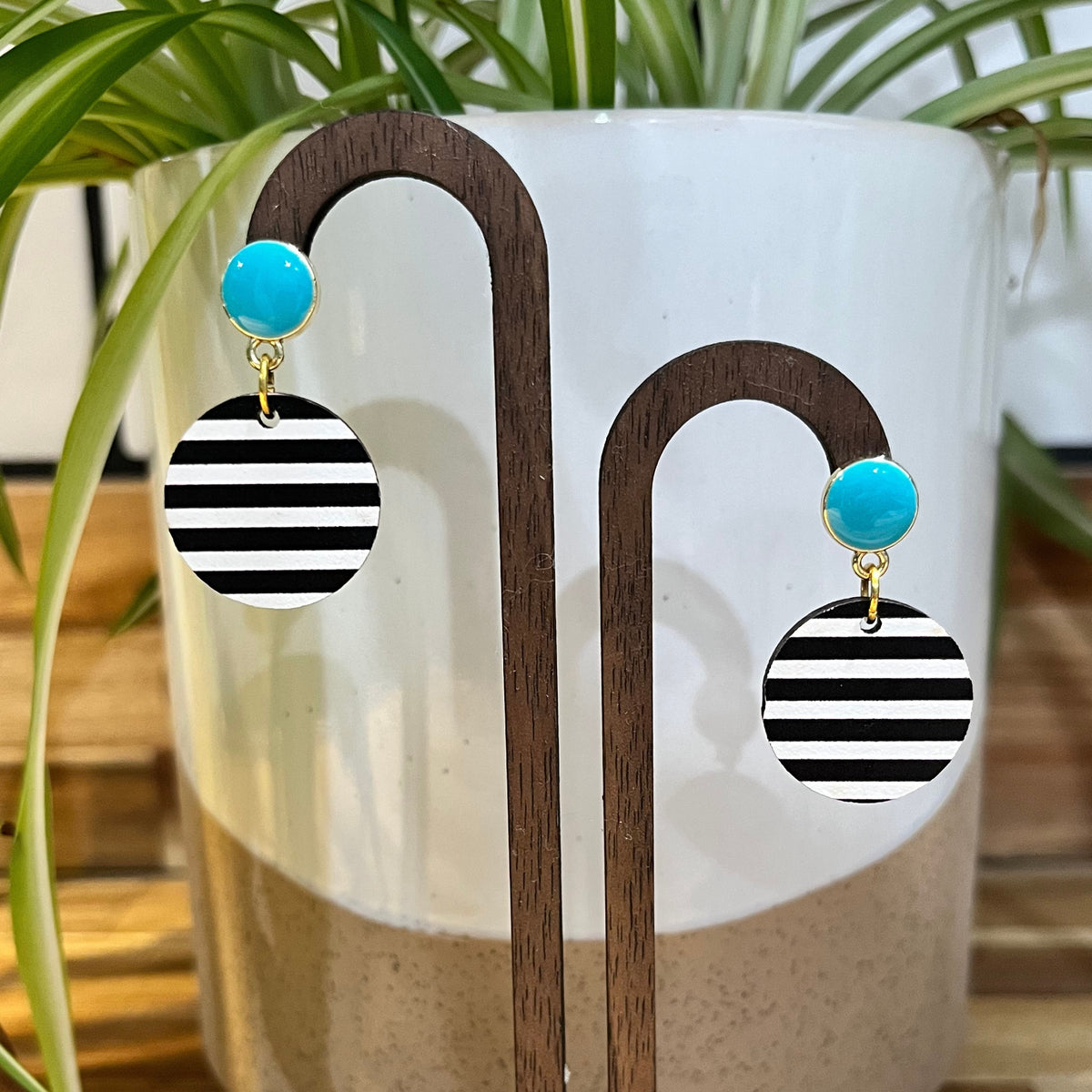 Black and white striped dangle earrings with turquoise blue round studs, displayed on a wooden stand in front of a white ceramic planter with green leaves.