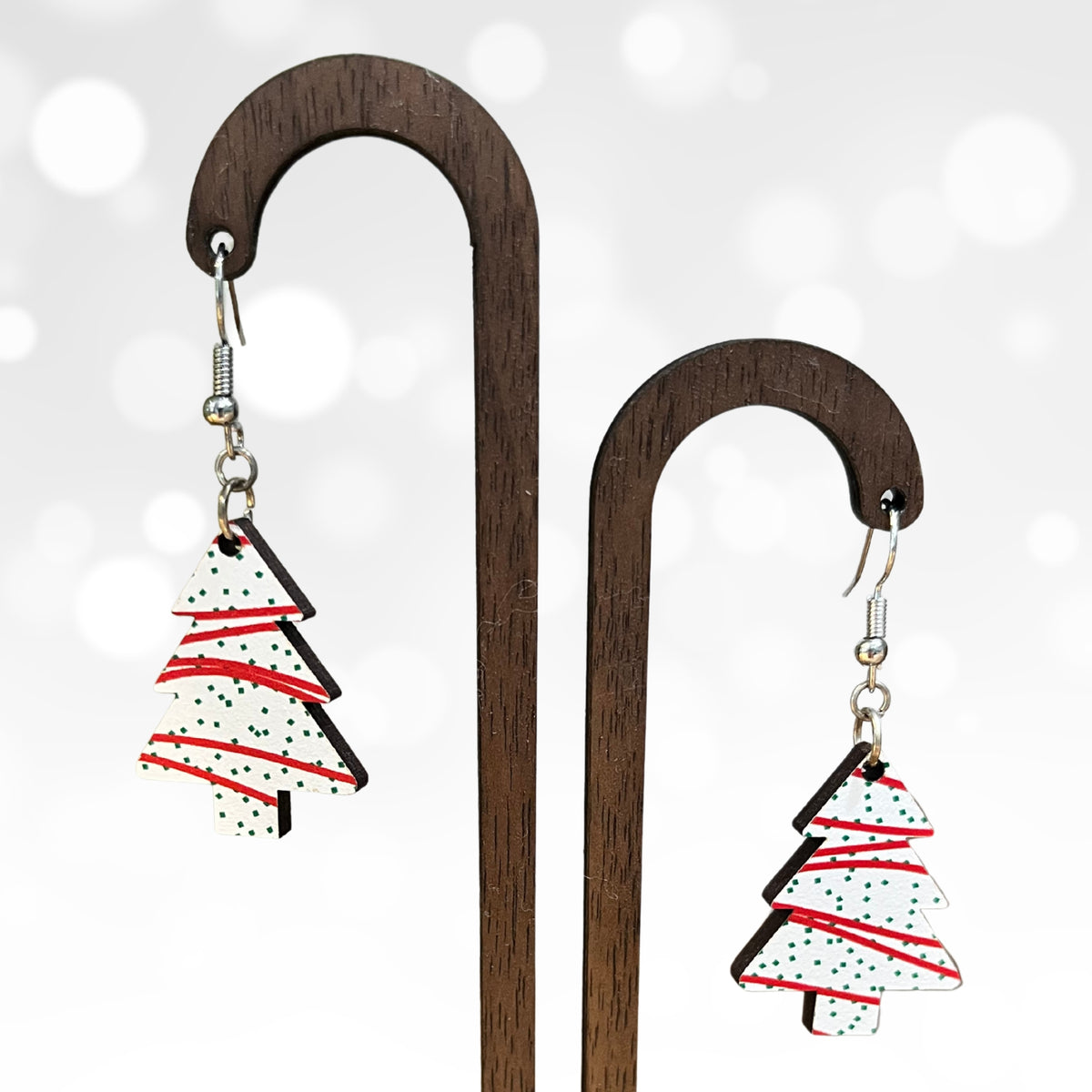 A pair of dangling earrings displayed on wooden hangers against a soft white bokeh background. Each earring has a silver fishhook ear wire and a small Christmas tree-shaped charm with a white background, red stripes, and black polka dots.