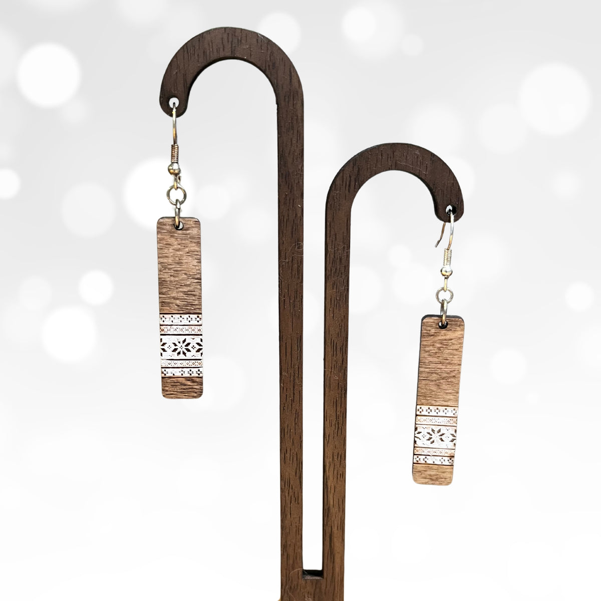 A pair of rectangular drop earrings with a natural wood grain finish and a white geometric pattern near the bottom, hanging from a dark wooden display stand against a soft white bokeh background.
