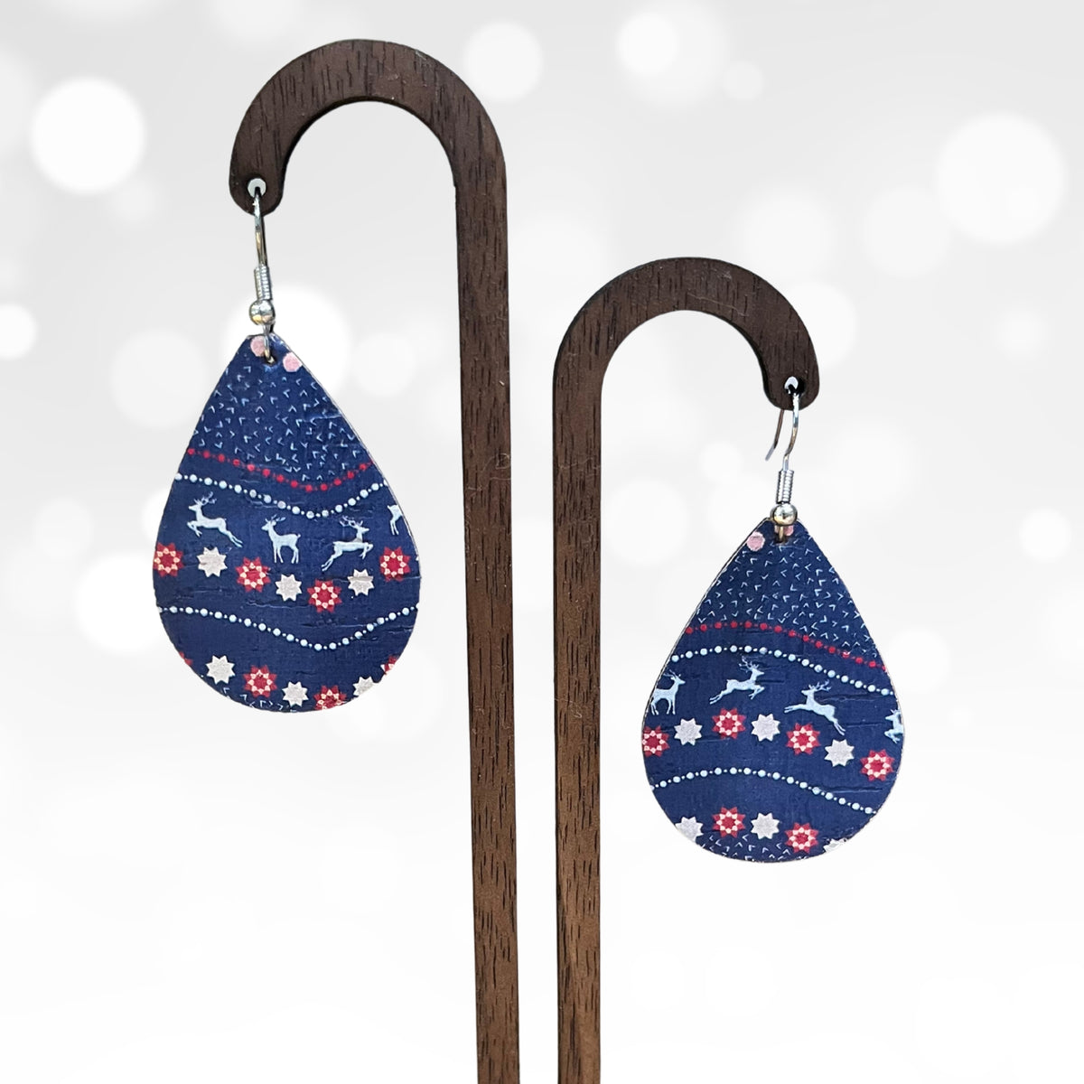 A pair of teardrop-shaped dangle earrings with a navy blue holiday pattern featuring white reindeer and red and white snowflake motifs, displayed on a wooden stand against a soft white bokeh background.