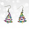 A pair of dangling earrings shaped like Christmas trees, filled with a multicolor confetti pattern, suspended from silver fishhook wires against a white background with soft bokeh light effects.