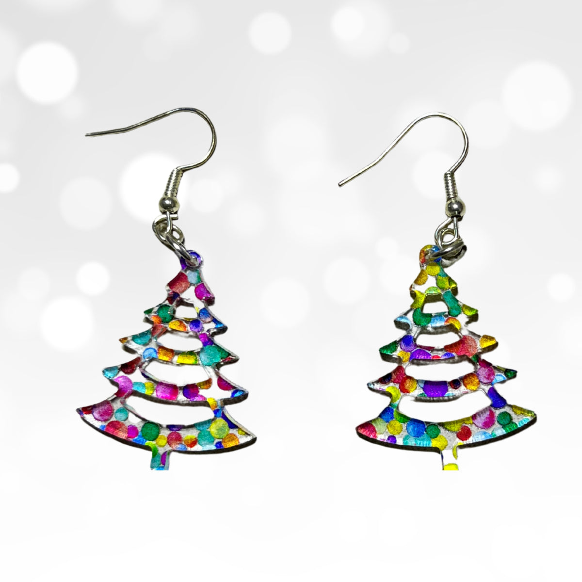 A pair of dangling earrings shaped like Christmas trees, filled with a multicolor confetti pattern, suspended from silver fishhook wires against a white background with soft bokeh light effects.