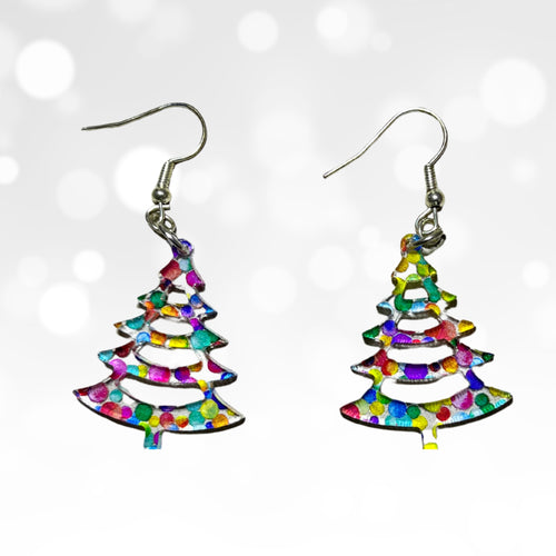 A pair of dangling earrings shaped like Christmas trees, filled with a multicolor confetti pattern, suspended from silver fishhook wires against a white background with soft bokeh light effects.