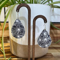 A pair of teardrop-shaped dangle earrings with a black and white floral starburst pattern, displayed on a wooden stand in front of a white ceramic planter with green leaves.
