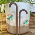A pair of gold-tone dangle earrings with round marbled resin pendants in teal, orange, and cream, displayed on a wooden stand in front of a white ceramic planter with green leaves.