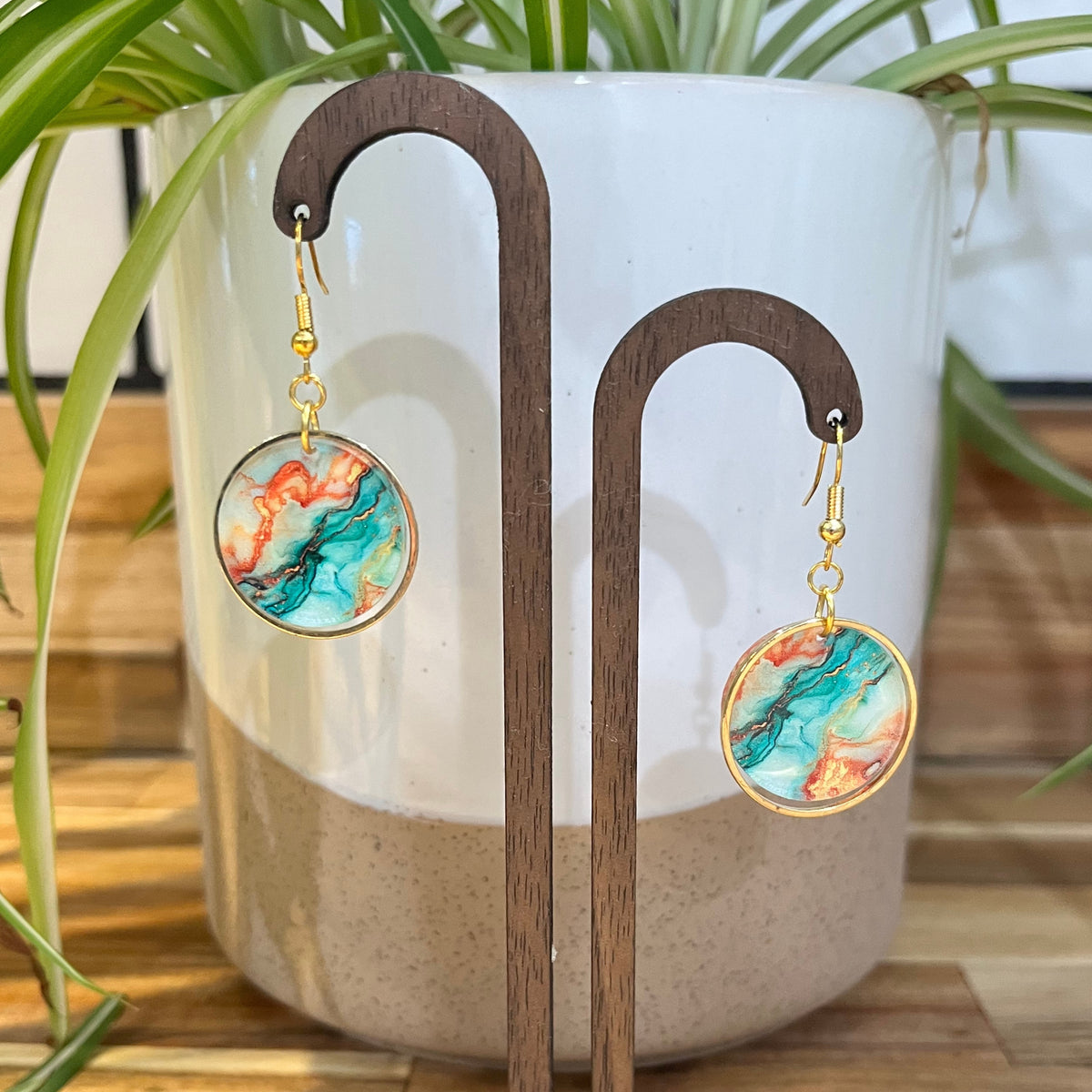 A pair of gold-tone dangle earrings with round marbled resin pendants in teal, orange, and cream, displayed on a wooden stand in front of a white ceramic planter with green leaves.