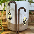 Leopard print drop earrings with gold-tone fishhook wires, displayed on a wooden stand in front of a white and beige ceramic planter with green plant leaves.