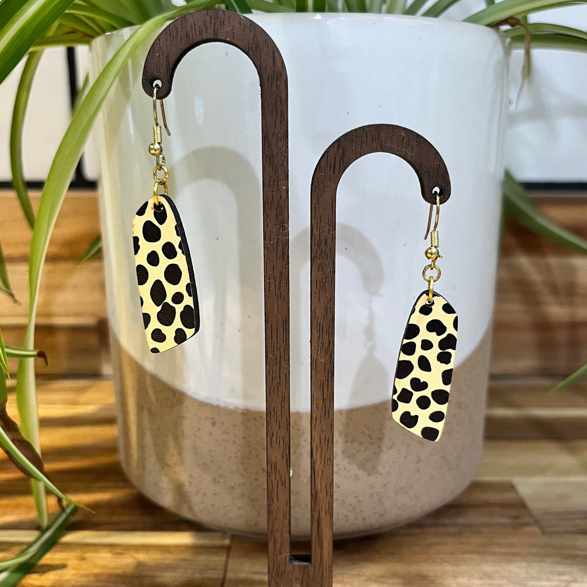 Leopard print drop earrings with gold-tone fishhook wires, displayed on a wooden stand in front of a white and beige ceramic planter with green plant leaves.