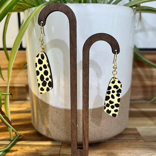 Leopard print drop earrings with gold-tone fishhook wires, displayed on a wooden stand in front of a white and beige ceramic planter with green plant leaves.