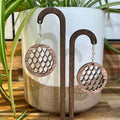 A pair of round wooden dangle earrings with a cutout honeycomb pattern, hanging from silver fishhook wires. They are displayed on a dark wood stand in front of a white ceramic planter with green plant leaves.