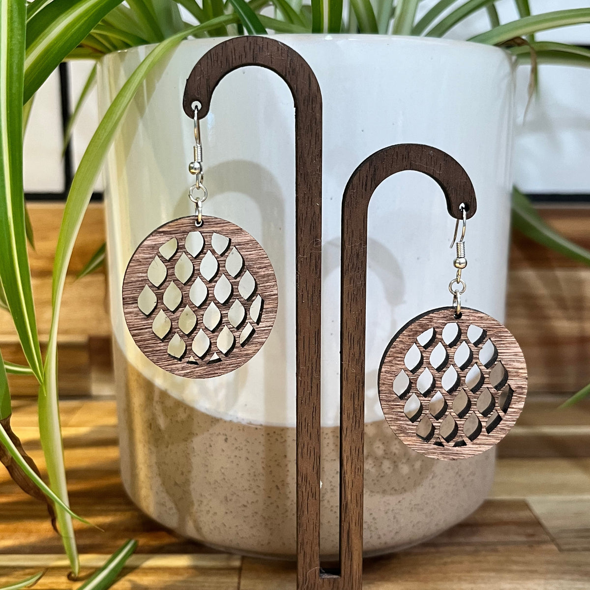 A pair of round wooden dangle earrings with a cutout honeycomb pattern, hanging from silver fishhook wires. They are displayed on a dark wood stand in front of a white ceramic planter with green plant leaves.