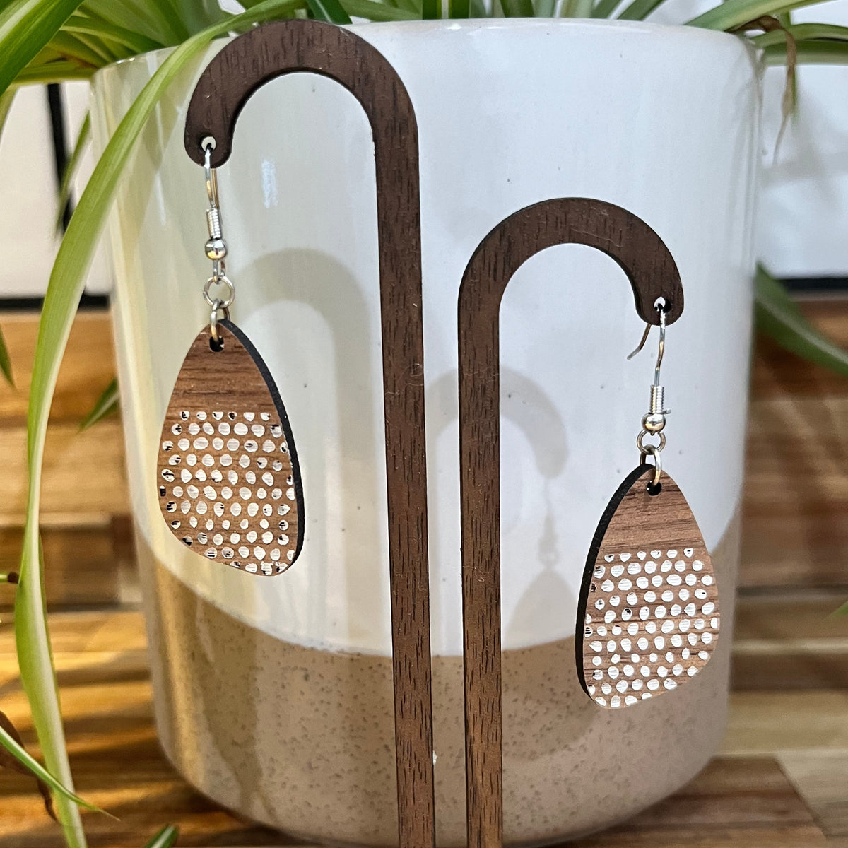 Wooden teardrop dangle earrings with a laser-cut honeycomb pattern, displayed on a wooden stand in front of a ceramic pot with green plant leaves.
