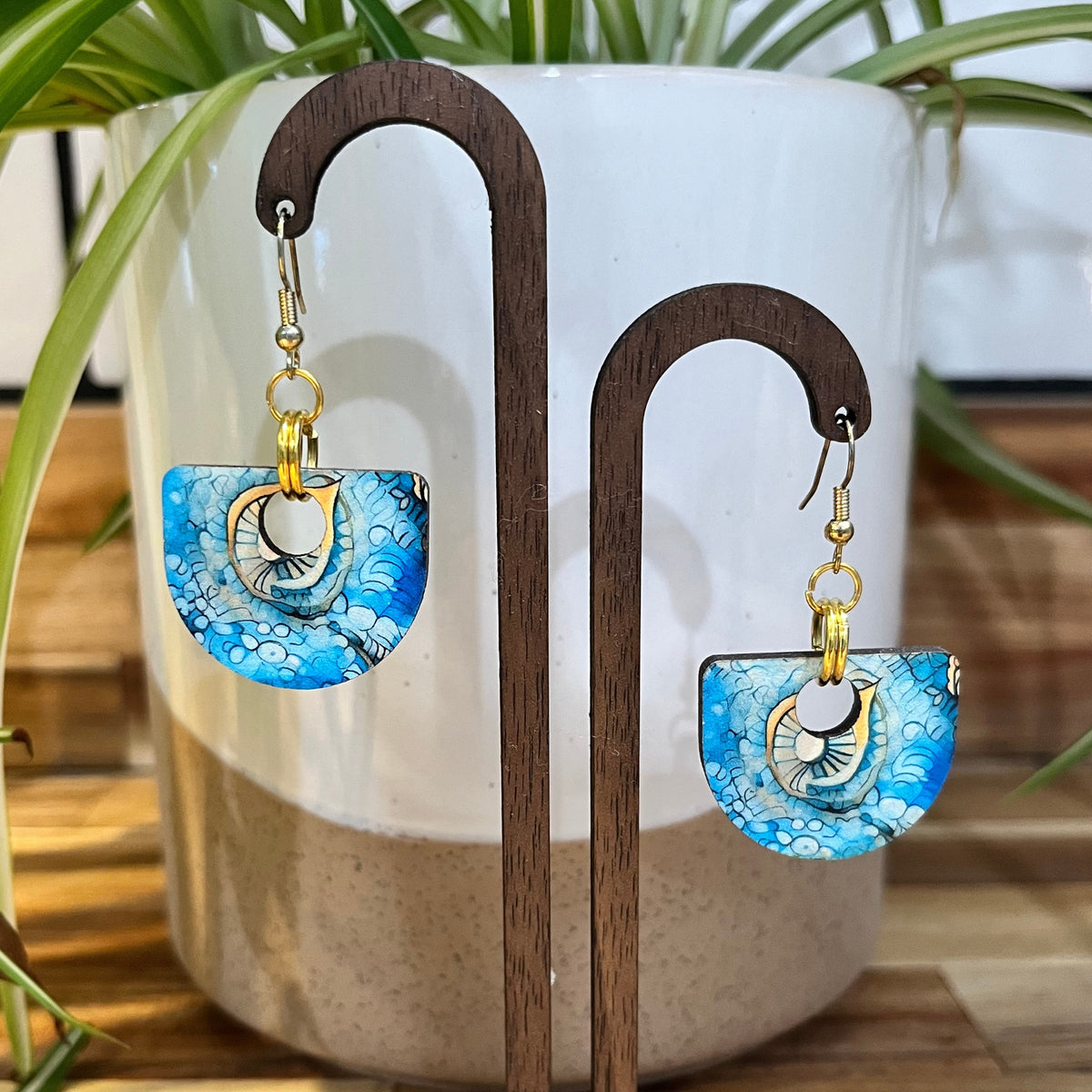 A pair of handmade dangle earrings displayed on a wooden stand in front of a white ceramic pot with green plant leaves. The earrings feature a blue and white patterned semi-circular drop with a gold-tone metal ring and a white circular center.