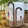 Colorful handmade beaded dangle earrings displayed on a wooden stand in front of a white ceramic planter with green plant leaves.