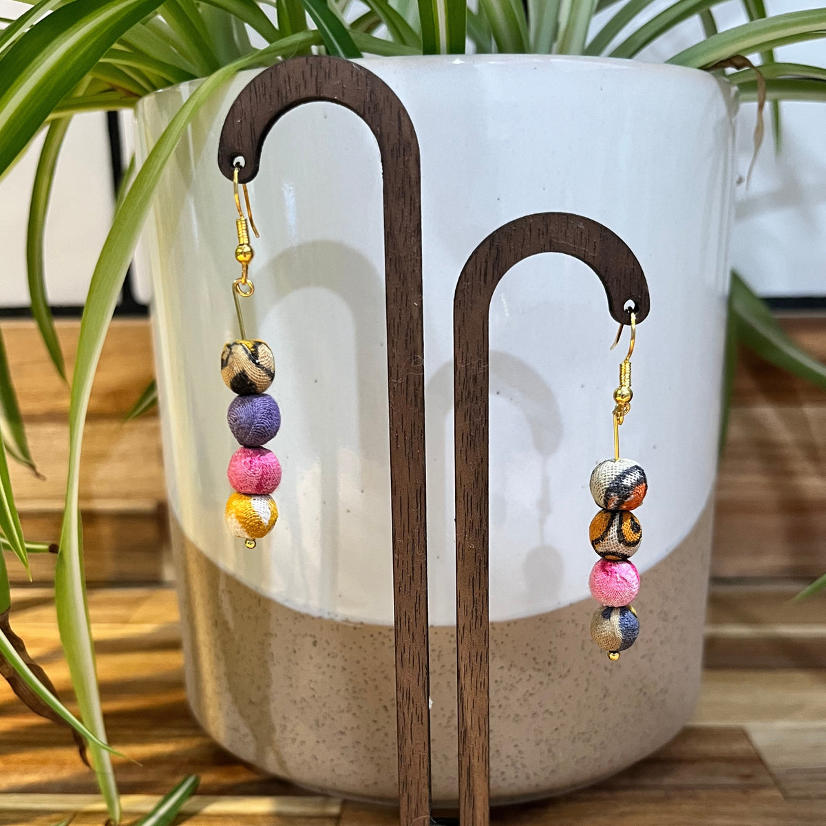 Colorful handmade beaded dangle earrings displayed on a wooden stand in front of a white ceramic planter with green plant leaves.