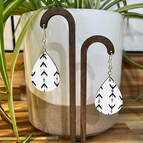 A pair of white teardrop-shaped dangle earrings with a black arrow pattern, hanging from a wooden display stand in front of a white ceramic planter with green plant leaves.