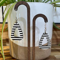 Black and white striped teardrop-shaped dangle earrings displayed on a wooden stand in front of a white ceramic planter with green plant leaves.