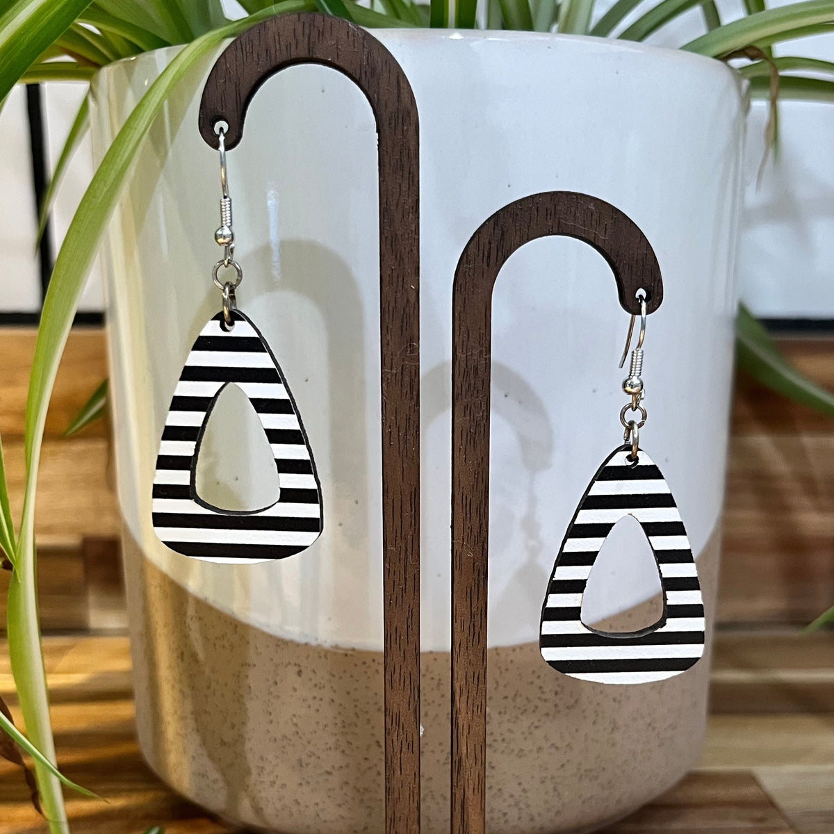 Black and white striped teardrop-shaped dangle earrings displayed on a wooden stand in front of a white ceramic planter with green plant leaves.