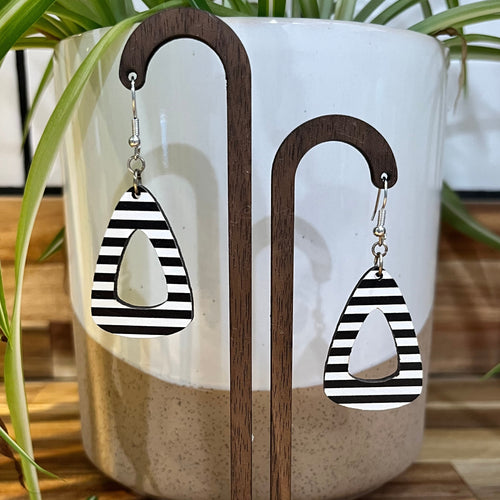 Black and white striped teardrop-shaped dangle earrings displayed on a wooden stand in front of a white ceramic planter with green plant leaves.
