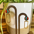 A pair of gold-tone dangle earrings with round red, orange, and turquoise marbled studs, displayed on a wooden stand in front of a white ceramic planter with green plant leaves.