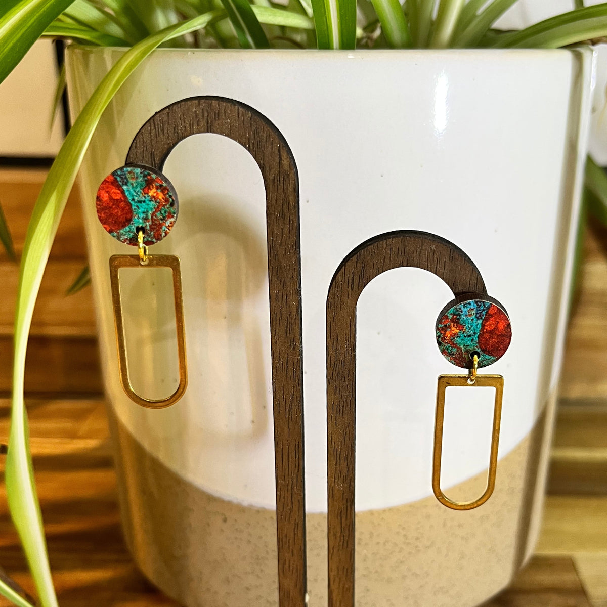 A pair of gold-tone dangle earrings with round red, orange, and turquoise marbled studs, displayed on a wooden stand in front of a white ceramic planter with green plant leaves.